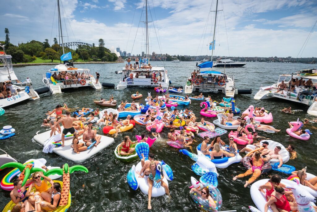 An image of a lively boat party on a sunny day. Multiple boats, including luxury yacht rentals Sydney, are decorated with flags and tied together in the water while numerous people on colorful inflatables enjoy music, swimming, and socializing. The background shows a city skyline and a bridge.