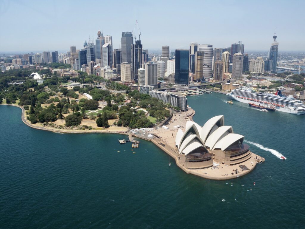 Aerial view of Sydney Opera House with its unique sail-like design, set against the backdrop of Sydney's cityscape and skyline. The Royal Botanic Garden is adjacent, with boats and a cruise ship visible in the harbor. Enjoy boat parties with The Yacht Social Club for an unforgettable experience on the water.