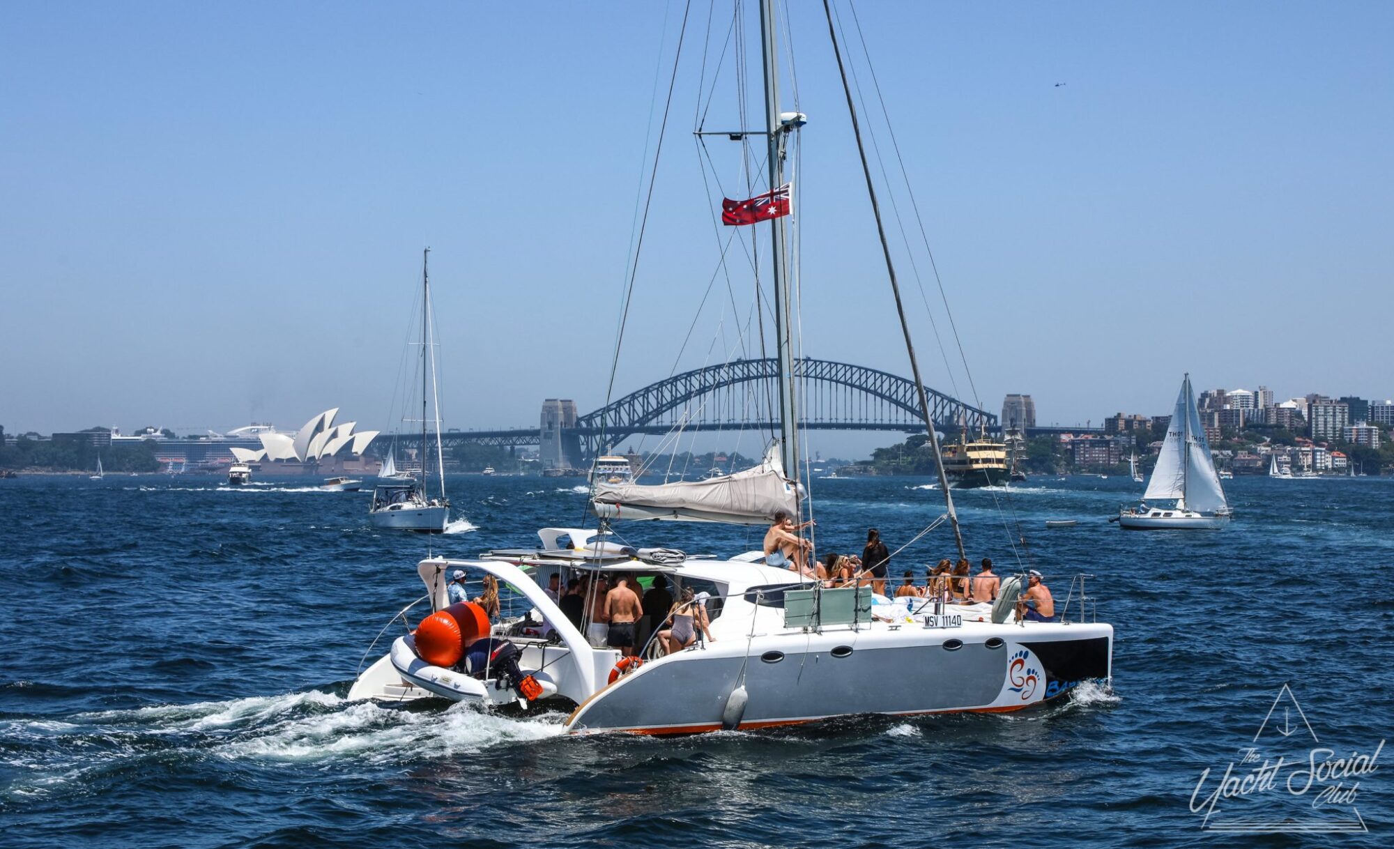 A catamaran party sails in a sunny harbor with the Sydney Opera House and Sydney Harbour Bridge visible in the background, along with other boats in the water.