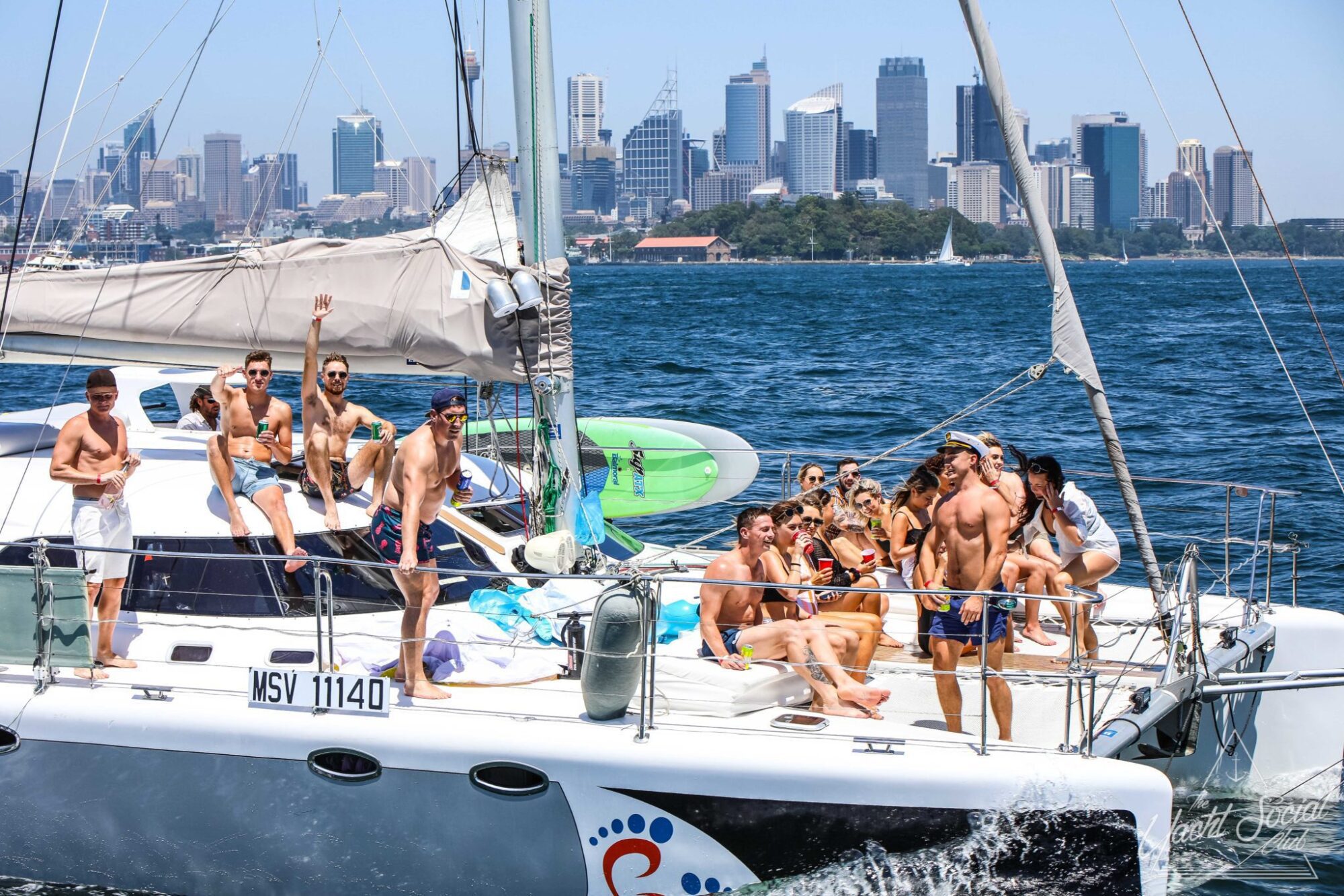 People are enjoying a sunny day on a sailboat with a city skyline in the background. The sailboat, identified as MSV 11140, is part of a Sydney boat party hire. Some are standing while others sit on the deck, taking in the beautiful views of Sydney Harbour.