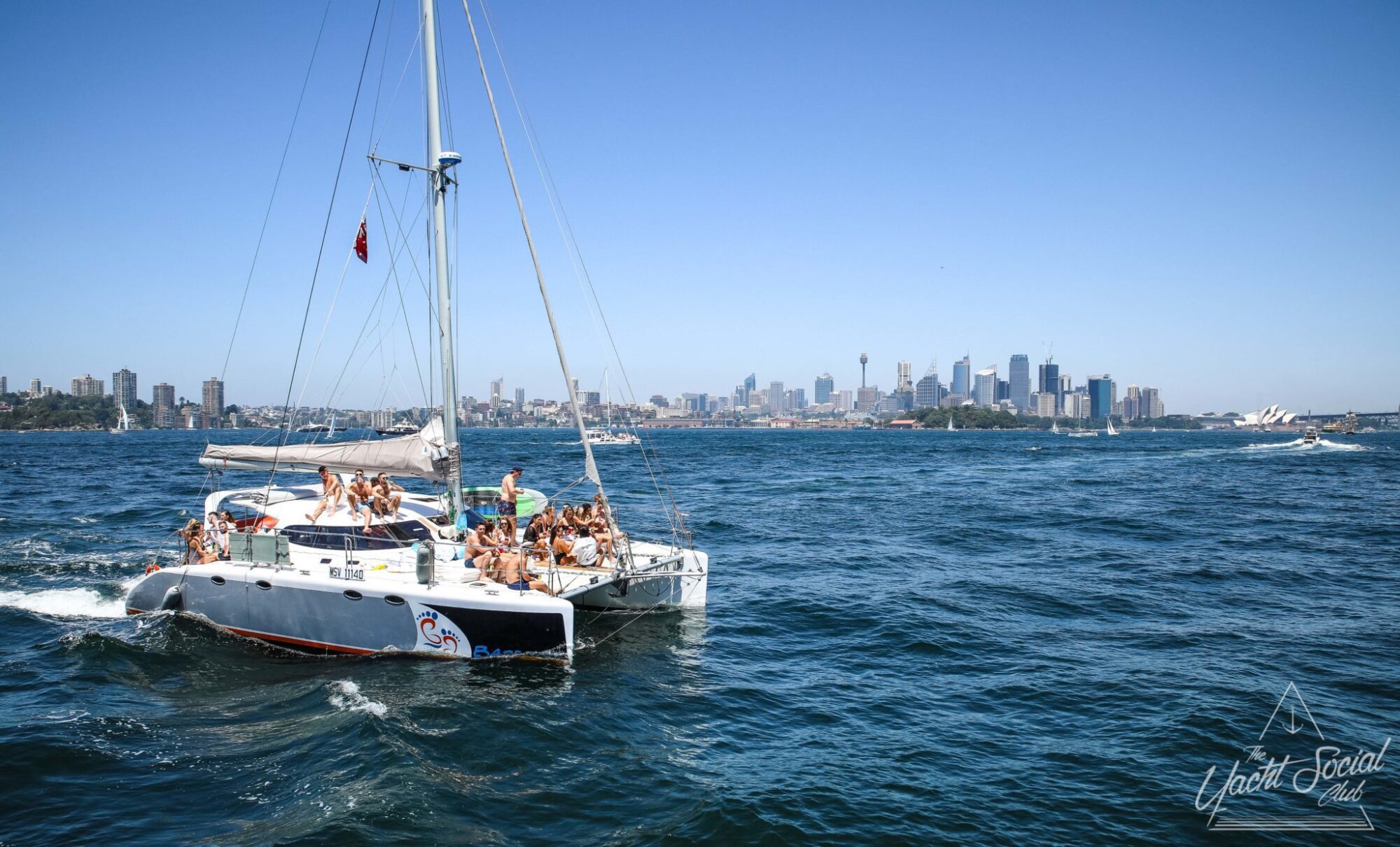 A luxurious catamaran with people onboard is sailing in a body of water with the city skyline and the Sydney Opera House in the background under a clear, blue sky. Ideal for DJ boat hire Sydney or catamaran party Sydney, this vessel offers an unforgettable experience.