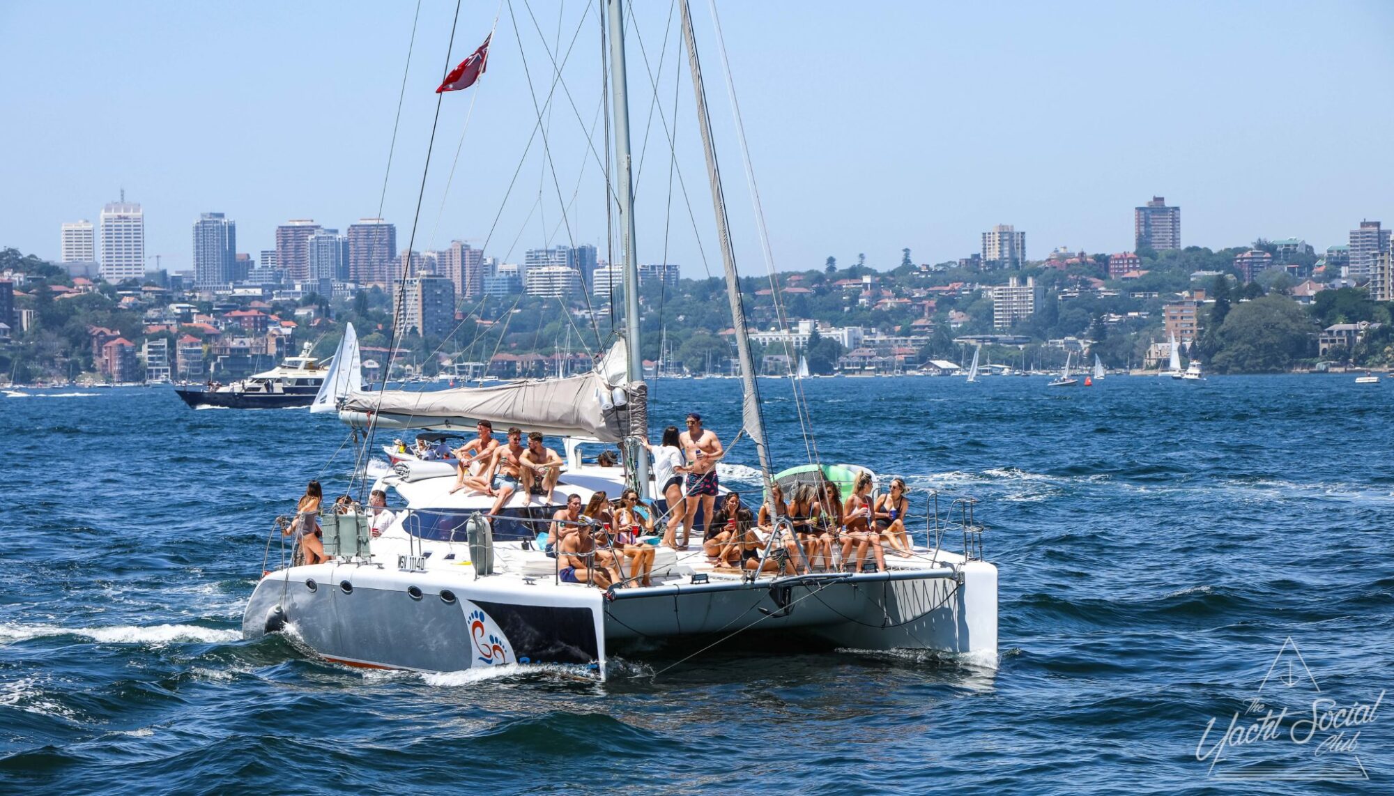 A catamaran with a group of people onboard sails on water with a city skyline and several other boats visible in the background, perfect for a corporate boat event in Sydney.