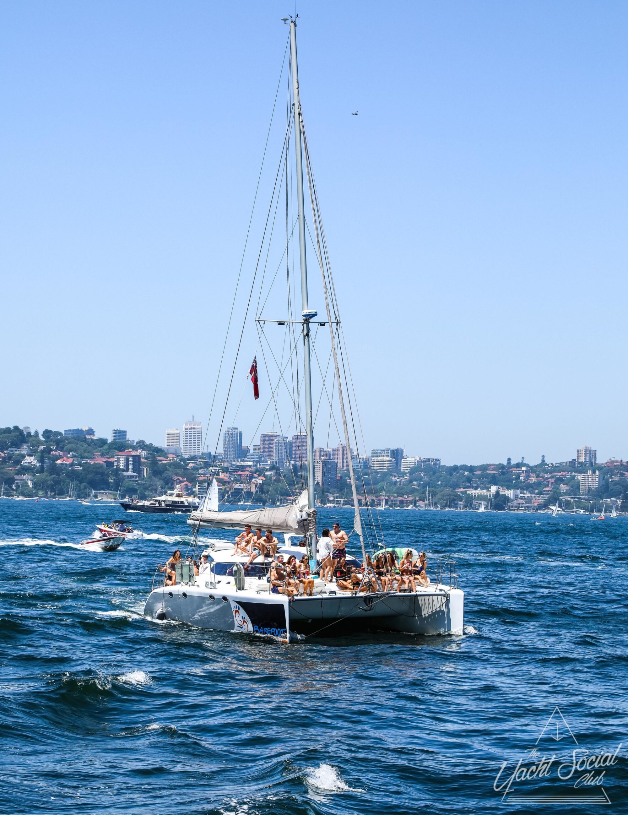 A large group of people are gathered on a sailing catamaran cruising the water, with city buildings visible in the background. Other boats can also be seen nearby, creating a lively atmosphere perfect for a Sydney boat party hire.