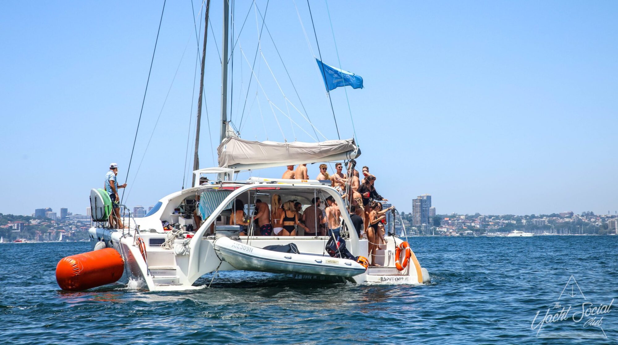 A group of people enjoying a day on the water aboard a catamaran sailboat, with a city skyline in the background and a blue flag on a sunny day. Perfect for corporate boat events Sydney or private yacht charter Sydney Harbour, it’s an ideal way to experience the stunning scenery and vibrant atmosphere.