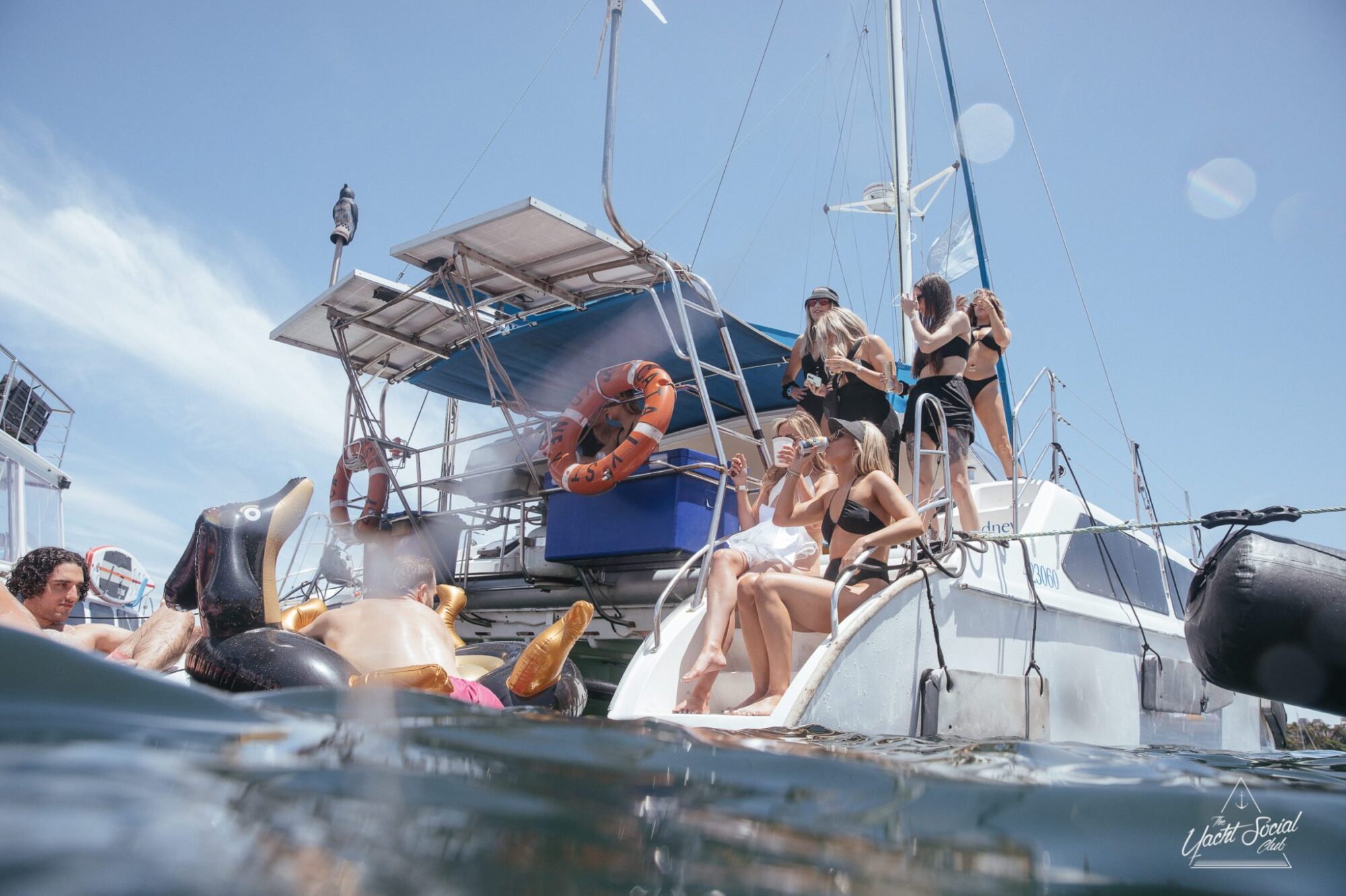 A group of people in swimwear are lounging and chatting on a sailboat, with inflatable float toys visible in the foreground. The boat is docked in calm water under a clear sky, setting the perfect scene for a DJ boat hire Sydney event.