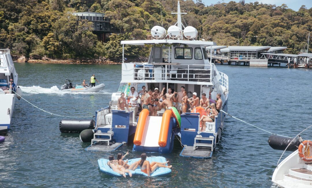 A lively group enjoys a party on a boat, with several individuals using an inflatable slide and floating devices in the water. A jetski and other boats are visible nearby, making it clear why Sydney boat party hire is so popular for corporate events and luxury yacht experiences.