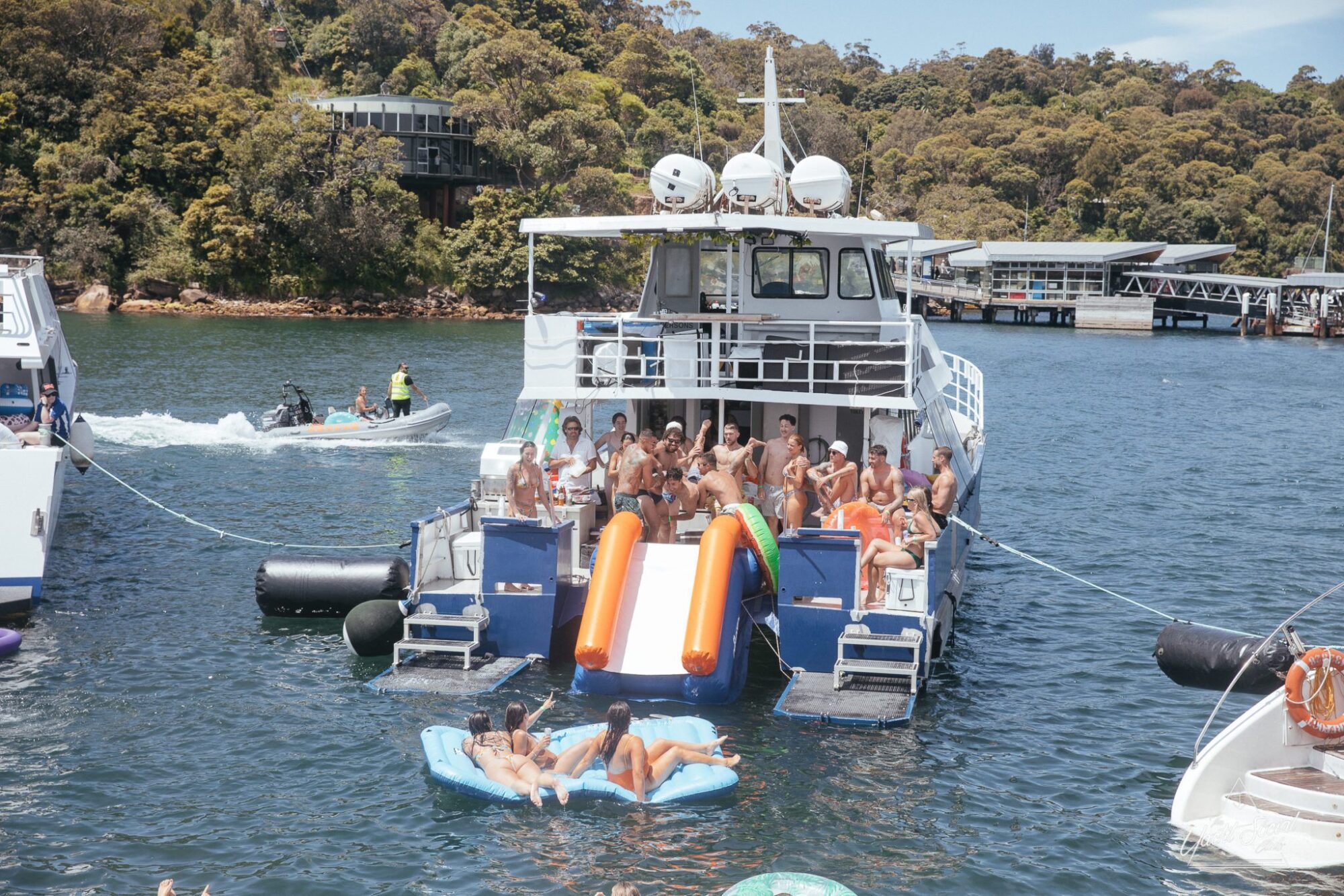 A lively group enjoys a party on a boat, with several individuals using an inflatable slide and floating devices in the water. A jetski and other boats are visible nearby, making it clear why Sydney boat party hire is so popular for corporate events and luxury yacht experiences.