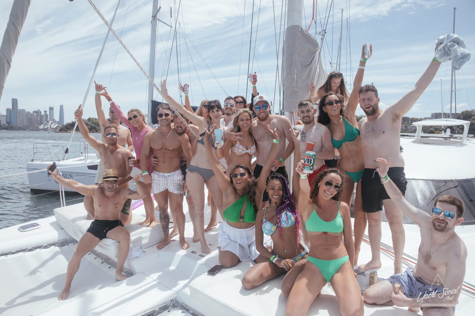 A group of people in swimwear posing and celebrating on a private yacht charter with sailboats and a city skyline in the background.