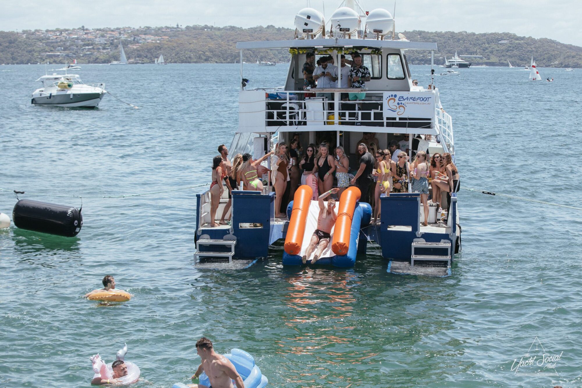 A luxury yacht hire in Sydney Harbour with numerous people onboard, some using slides into the water. Additional people are floating and swimming nearby. The background includes other boats and a distant shoreline.