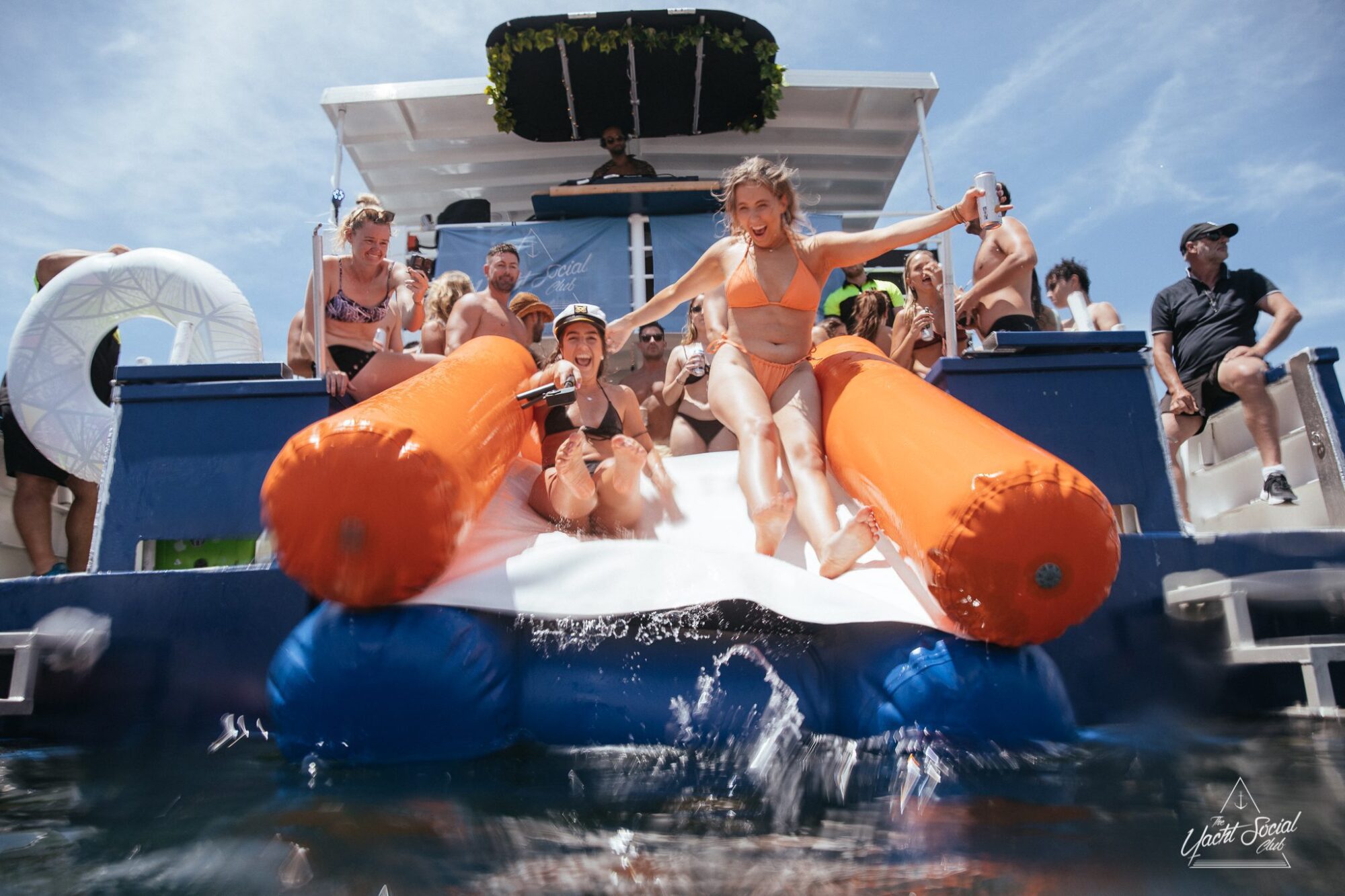 A group of people enjoying a sunny day on a boat with two women sliding down an inflatable slide into the water during a Sydney boat party hire event.