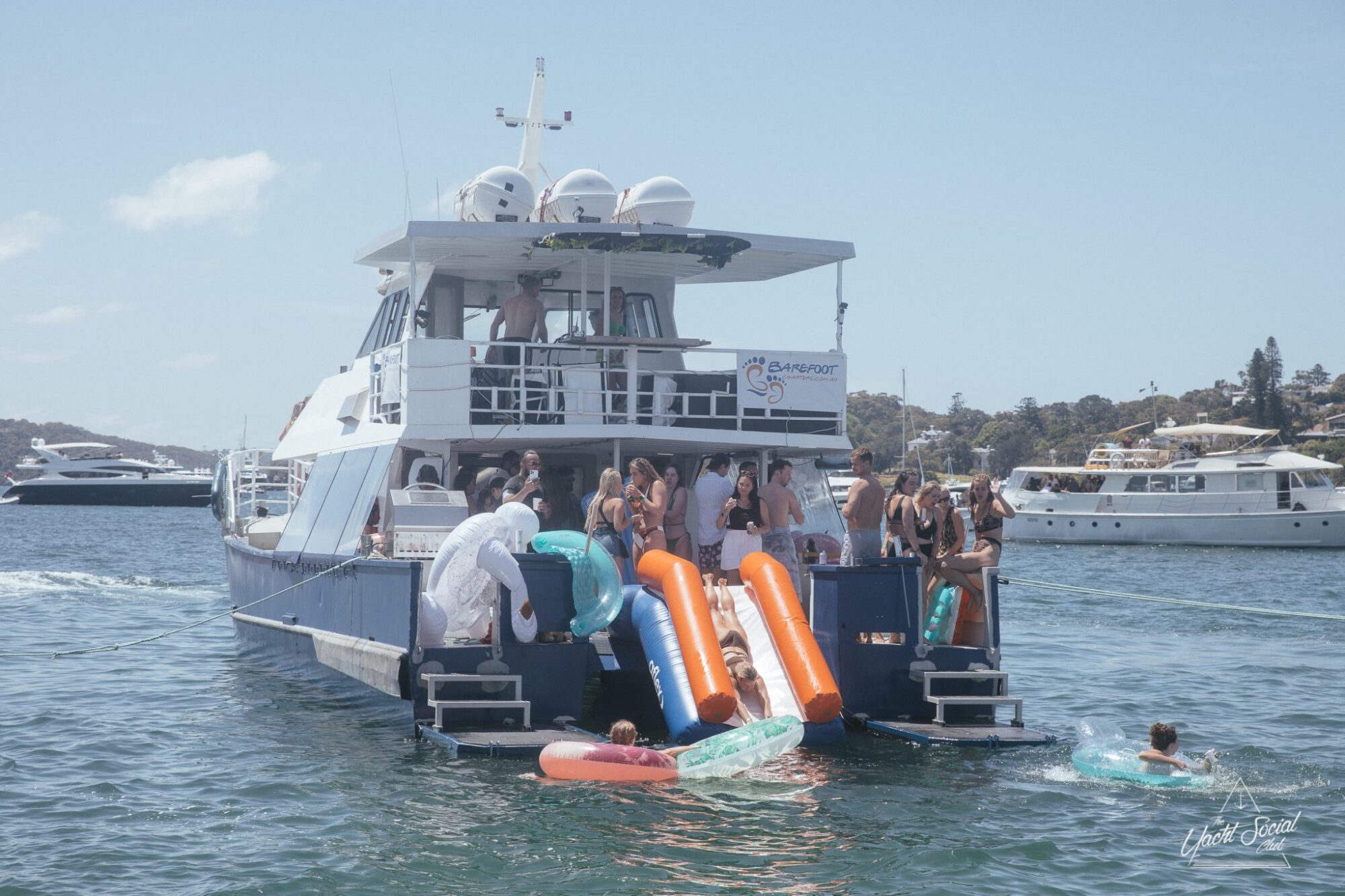 A group of people are enjoying themselves on a private yacht charter in Sydney Harbour with slides and inflatables, anchored in a sunny harbor surrounded by other boats.