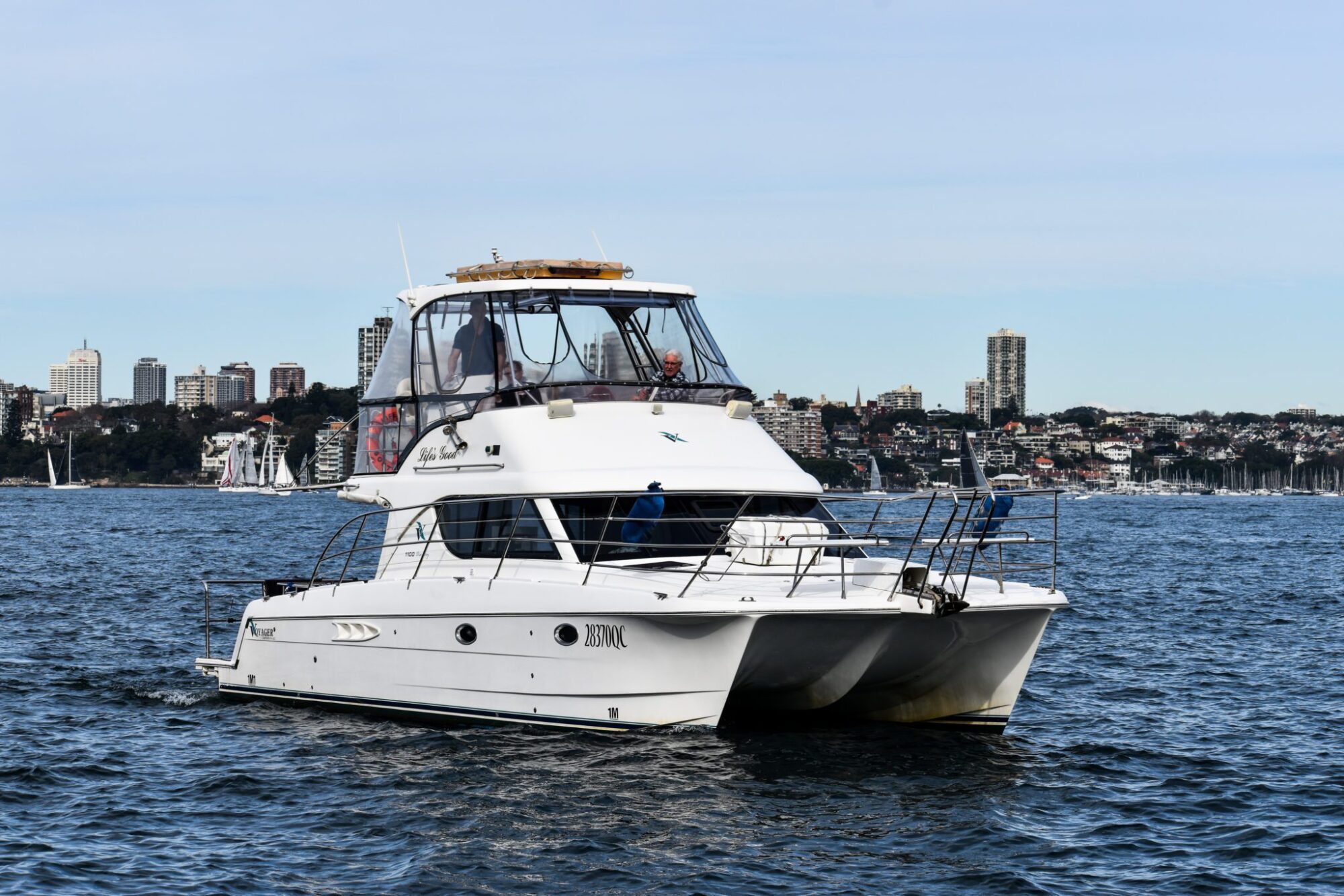 A white motor yacht is sailing on a calm body of water with a cityscape of tall buildings and trees visible in the background. The sky is clear, perfect for a corporate boat event or private yacht charter on Sydney Harbour.