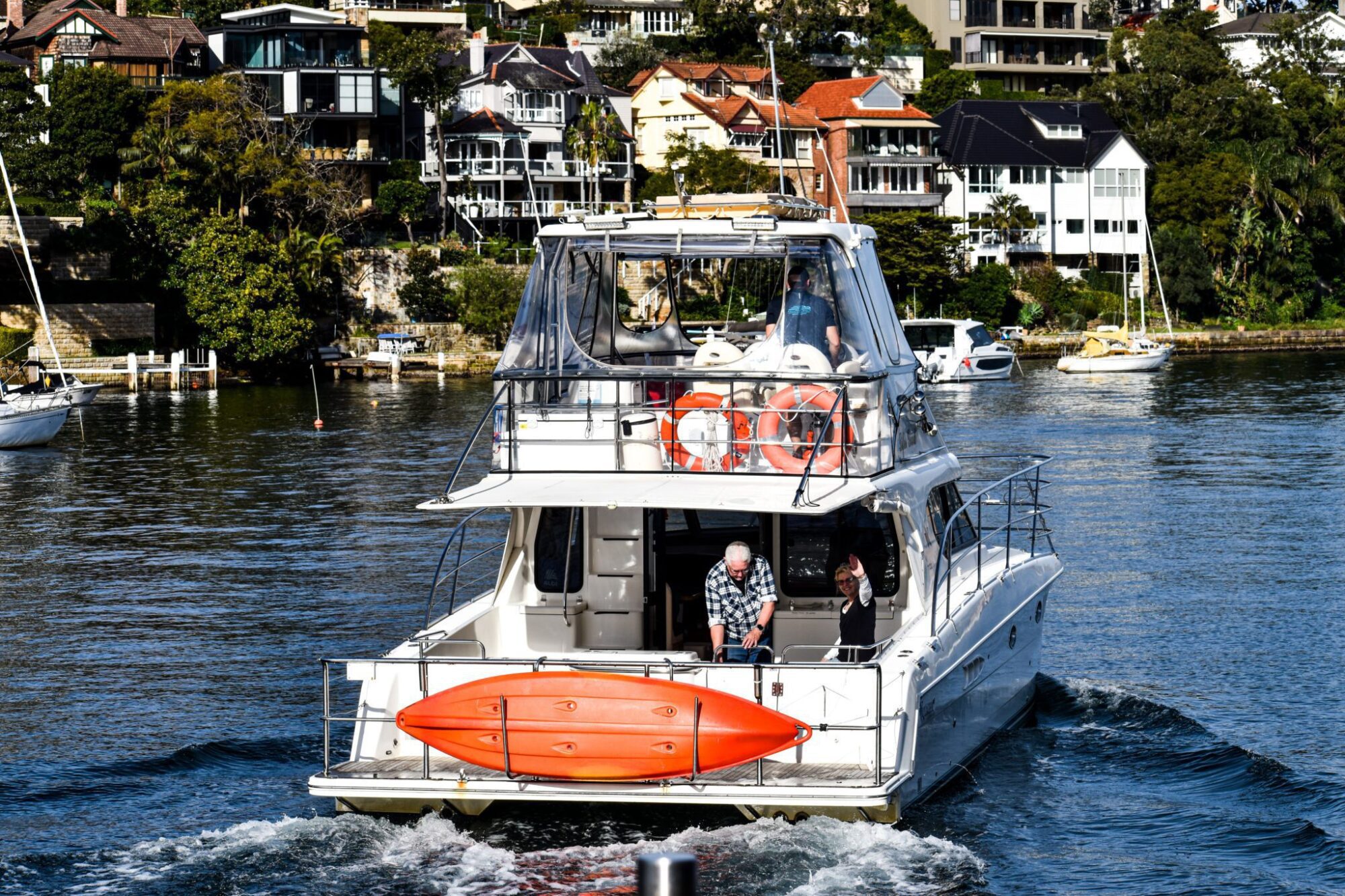 A motorboat with people, including a man in a checkered shirt, travels through a waterway, passing by houses on the shore. An orange kayak is mounted on the back of the boat. Perfect for corporate boat events in Sydney or even hiring for your own Sydney boat party adventure!