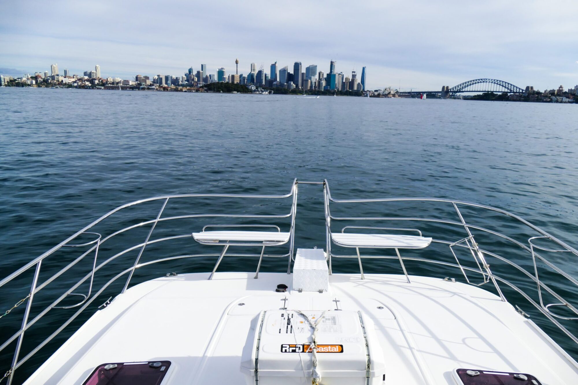 A view of a city skyline with prominent skyscrapers and a bridge, seen from the bow of a luxury yacht floating on a calm body of water.