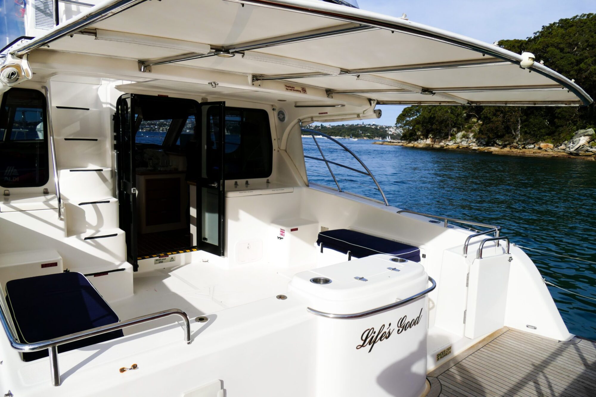 The image shows the rear deck of a boat named "Life's Good." The deck has seating areas with navy cushions, a small table, and an open doorway leading inside. The boat, perfect for a private yacht charter in Sydney Harbour, is on calm waters near a forested shoreline.