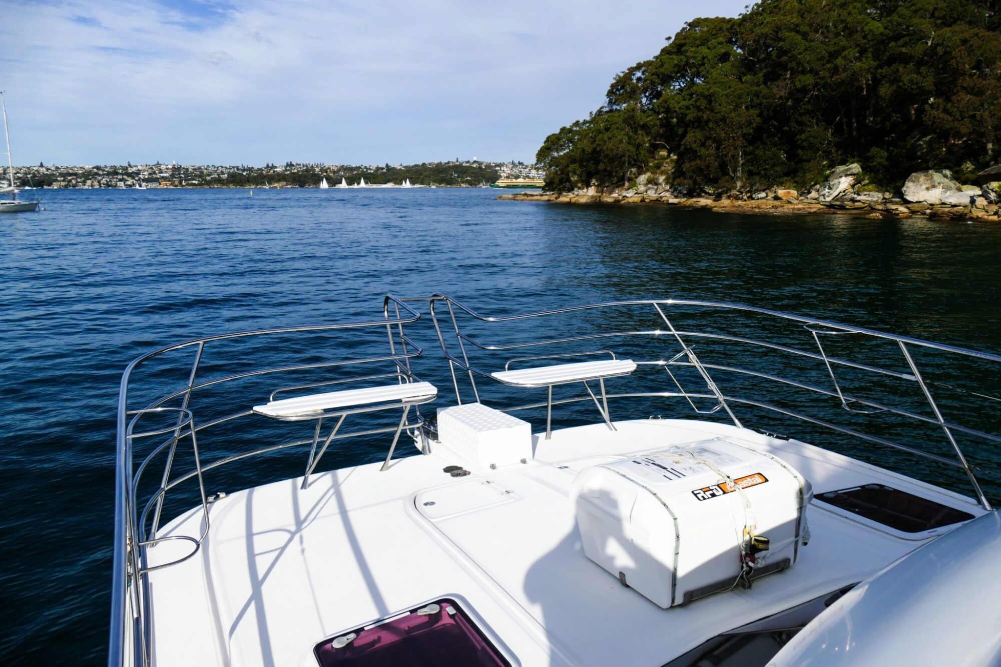 View from the front deck of a boat on a calm body of water with a tree-lined shoreline and distant sailboats in the background, perfect for a luxury yacht hire Sydney adventure.