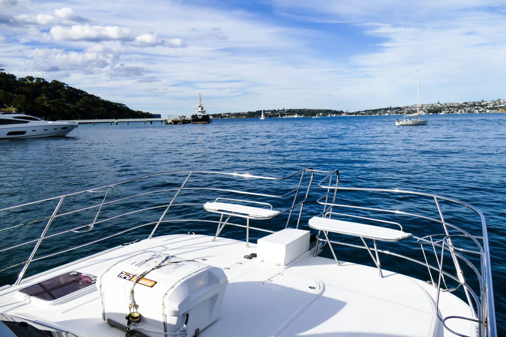 Enjoy the view from the deck of a boat showing a calm body of water with several other boats and a hilly coastline in the background under a partly cloudy sky. Perfect for private yacht charter Sydney Harbour or corporate boat events.