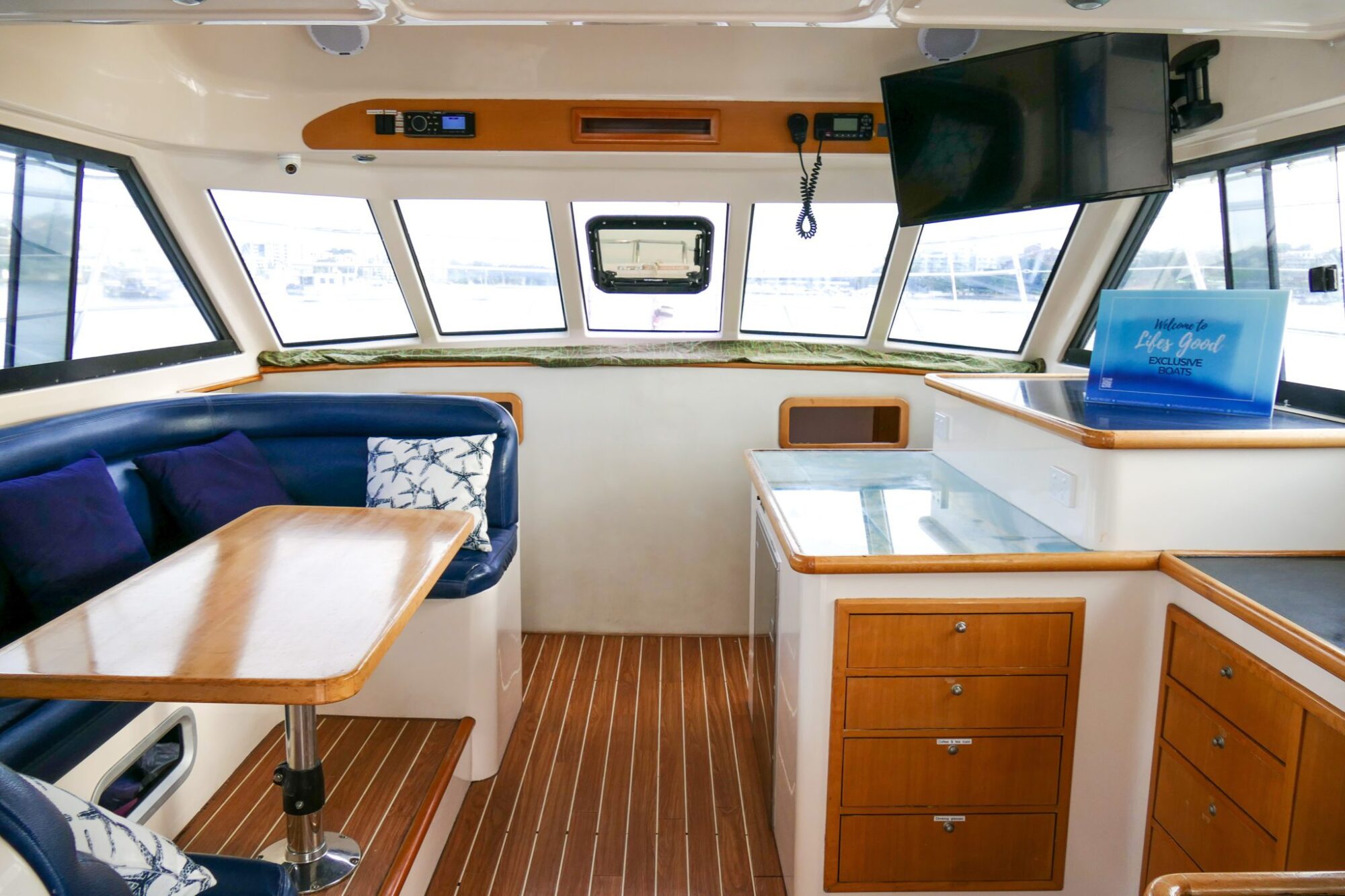 Interior of a boat featuring a seating area with a wooden table, blue cushions, and a small kitchen with cabinets. A mounted TV completes the setup. Windows provide stunning views of the water outside, perfect for a luxury yacht hire Sydney experience.