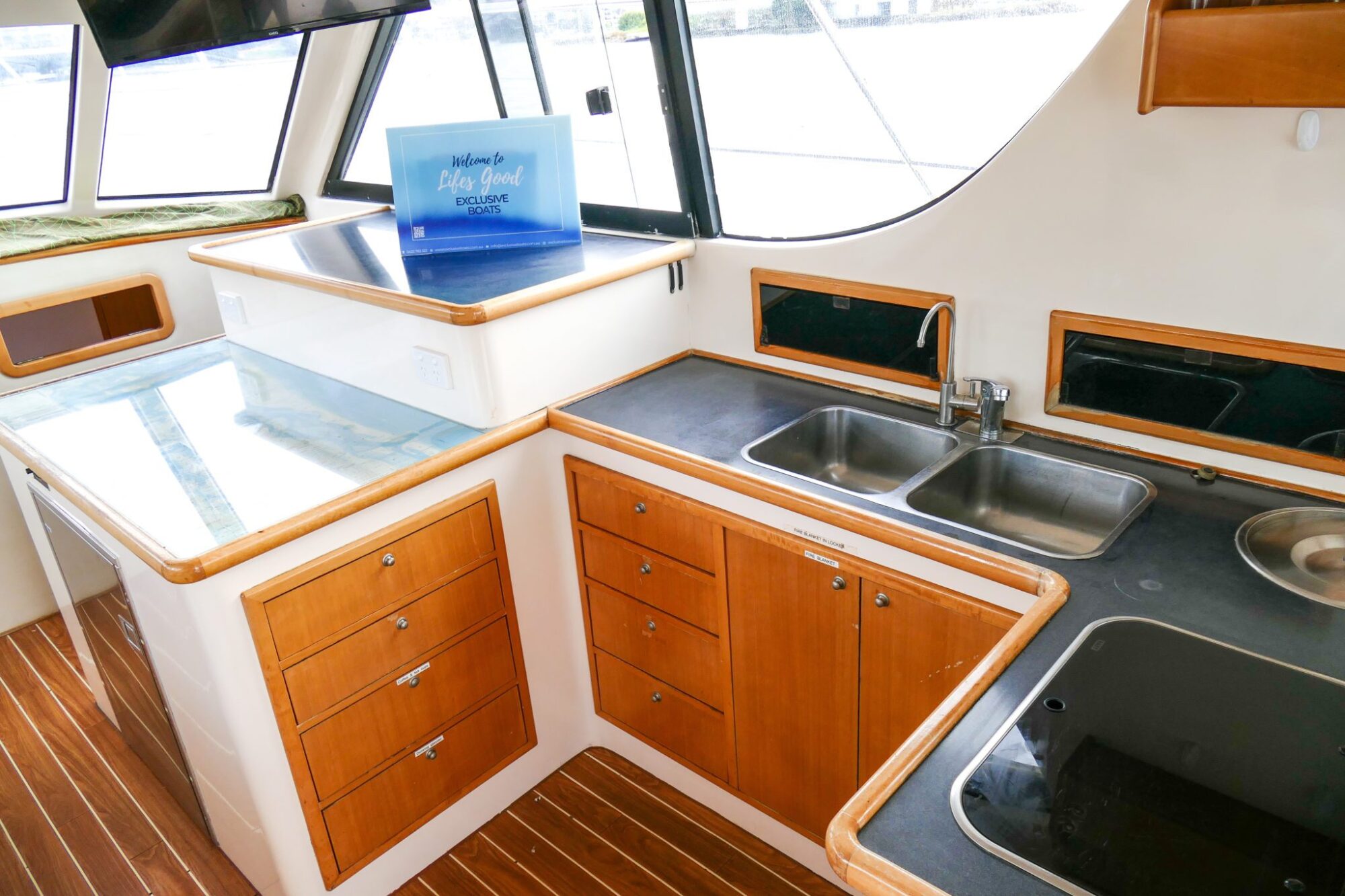 Interior of a boat kitchen featuring wooden cabinetry, a double sink, stovetop, and countertop with a sign reading "Life is good" propped on the counter. Natural light filters through the windows. Perfect for those considering private yacht charter Sydney Harbour trips.