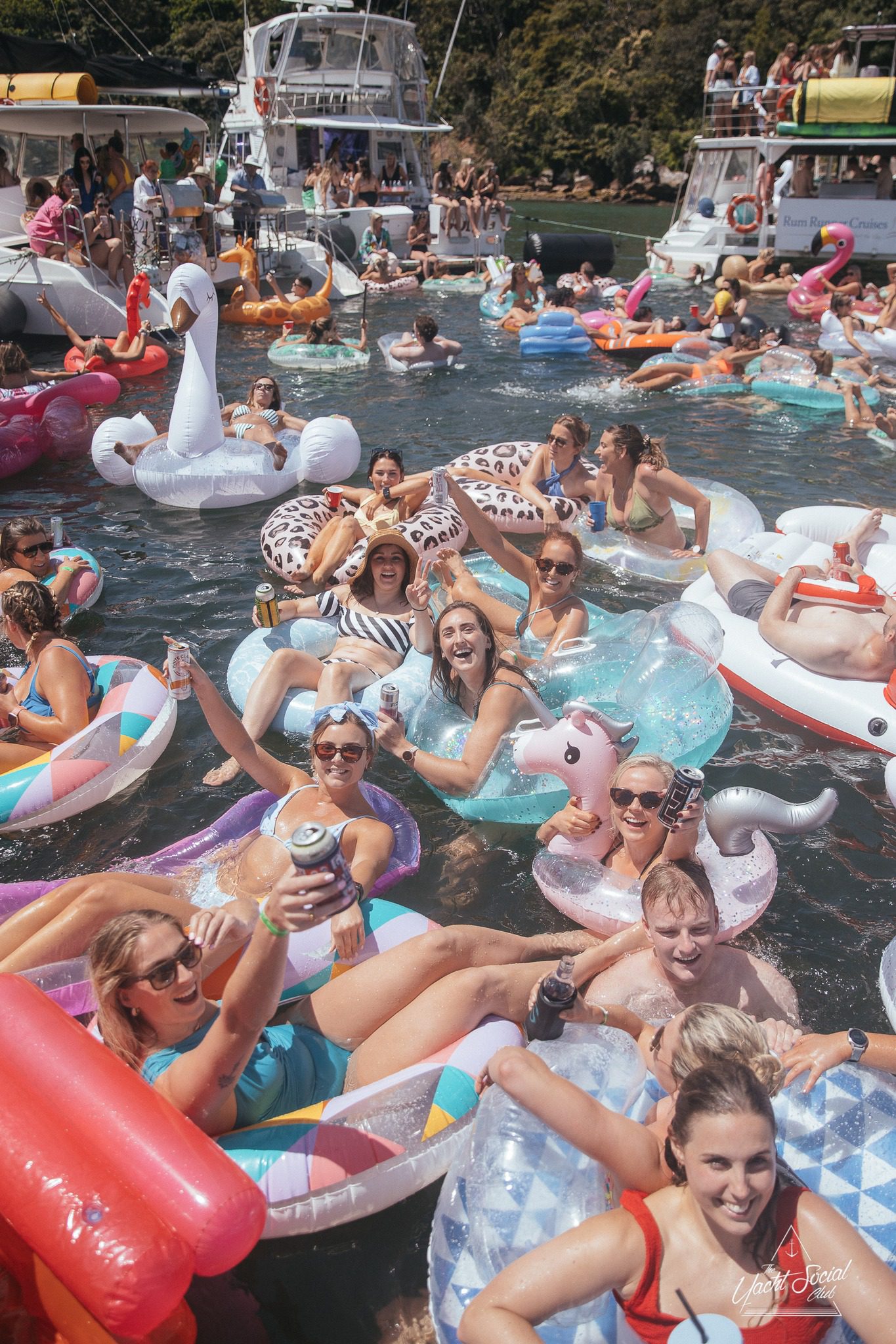 A group of people enjoying a sunny day on a lake, lounging on various inflatable pool floats, with boats and other participants in the background, reminiscent of a Sydney boat party hire.