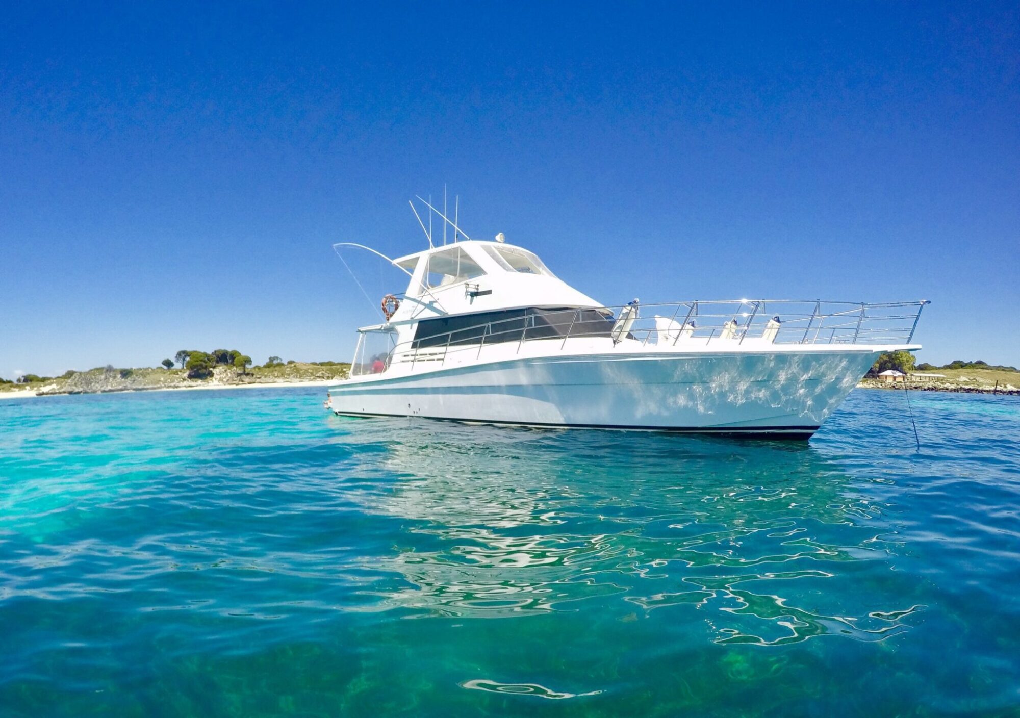 White motor yacht anchored in clear blue water with a coastline and clear blue sky in the background, perfect for a Sydney boat party hire.