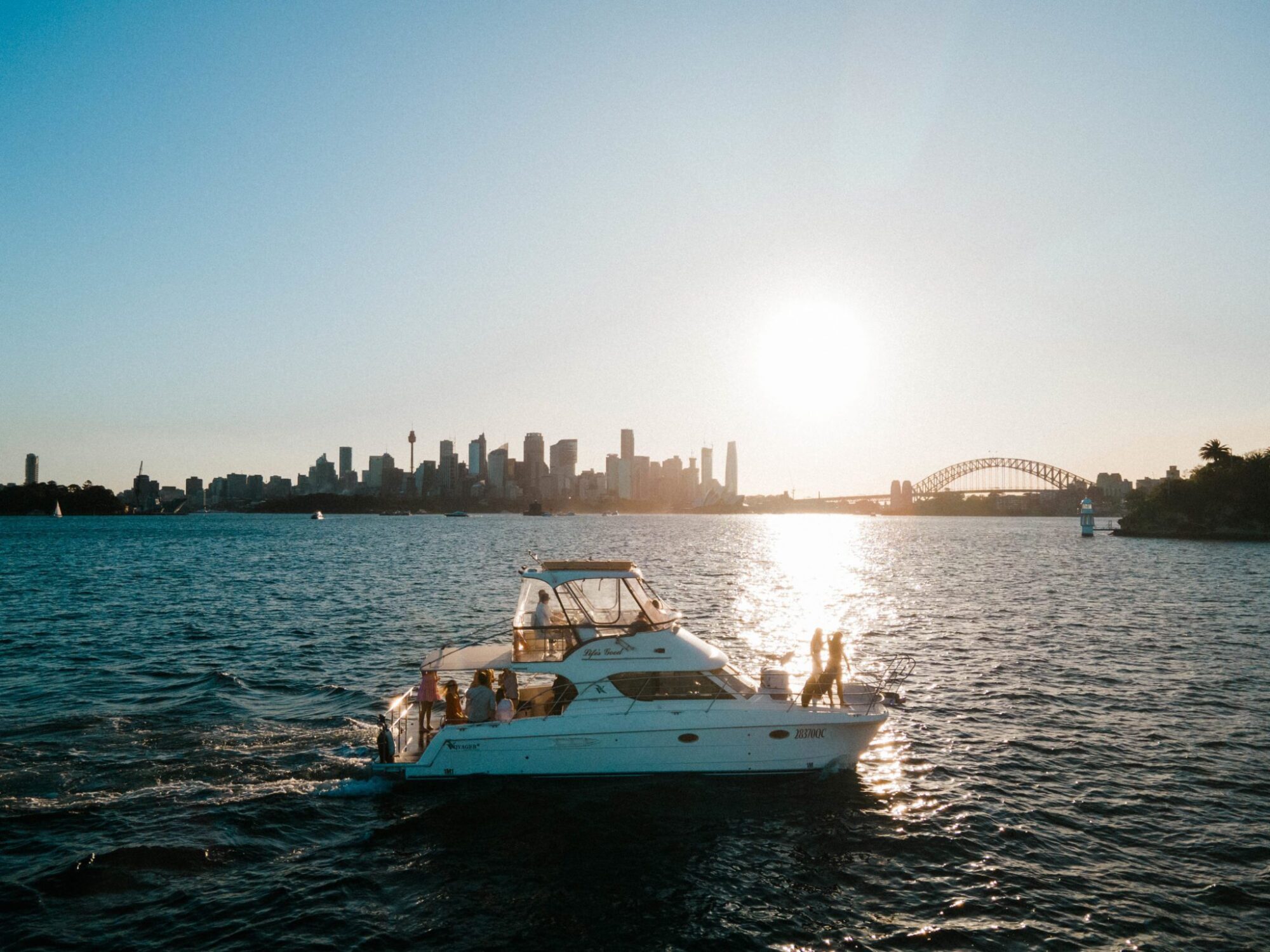 A luxury yacht hire Sydney sails on a body of water during sunset with a city skyline and a bridge visible in the background.