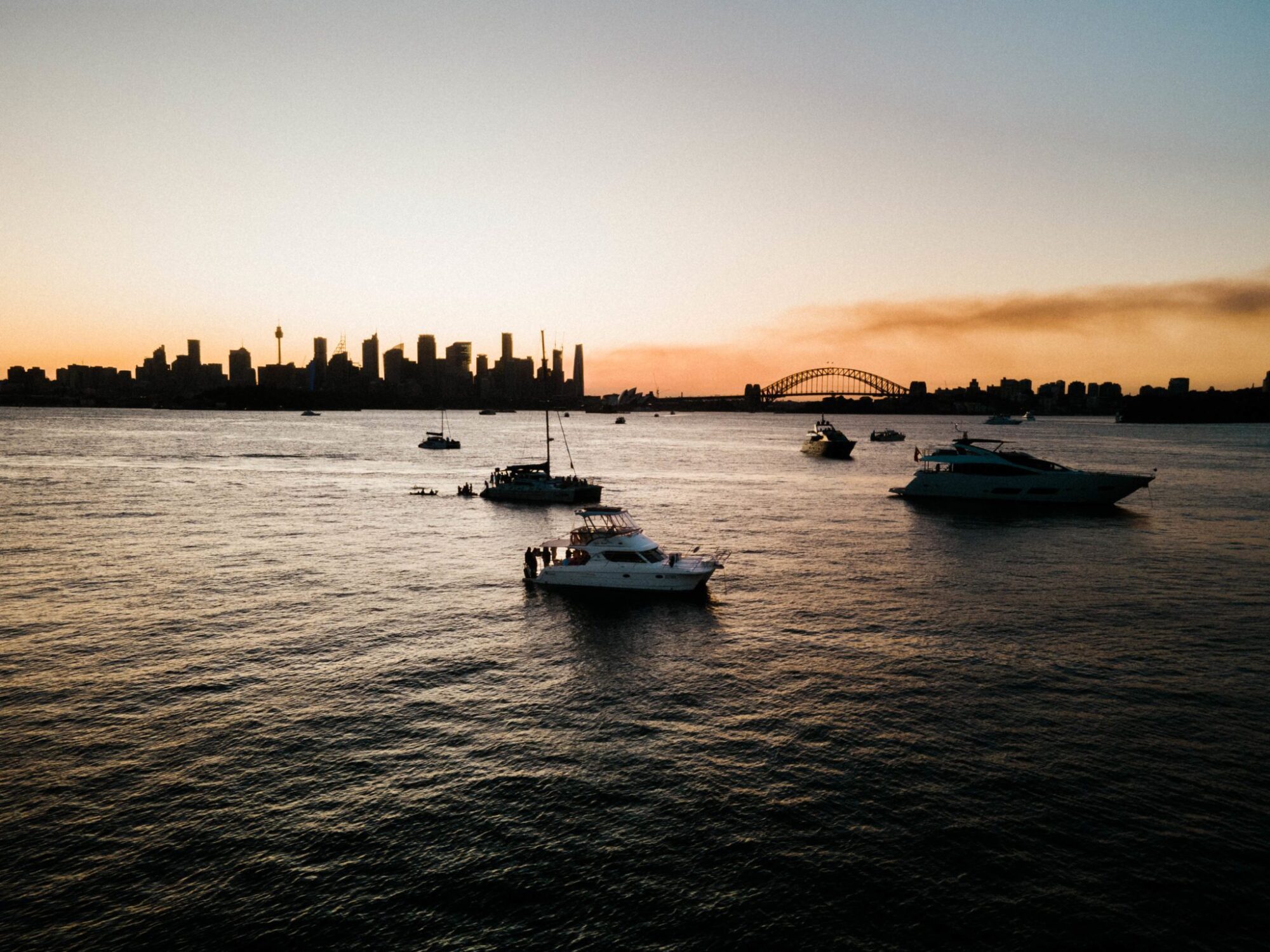 Boats float on a calm body of water with a city skyline and a bridge in the background at dusk, perfect for luxury yacht hire Sydney.