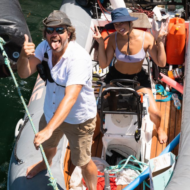 Two people on a small boat; one man in a white shirt and hat is making a silly face and gesturing while a woman in a bikini and hat behind him stands by the steering wheel, making a peace sign. Perfect vibes for your next Sydney boat party hire or corporate boat event!