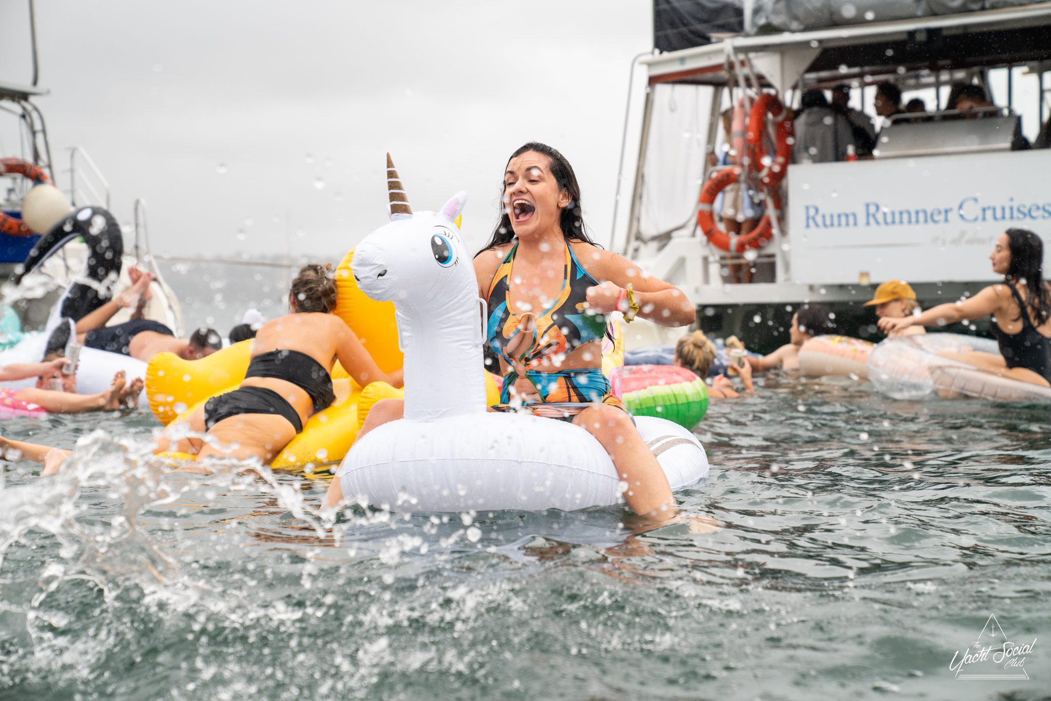 A woman wearing a swimsuit rides an inflatable unicorn while splashing in water among other people and inflatables near a boat labeled "Rum Runner Cruises," enjoying a lively Catamaran party Sydney.