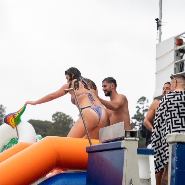 People are on a luxury yacht, with some standing and one person preparing to slide down an orange water slide into the water near an inflatable unicorn.
