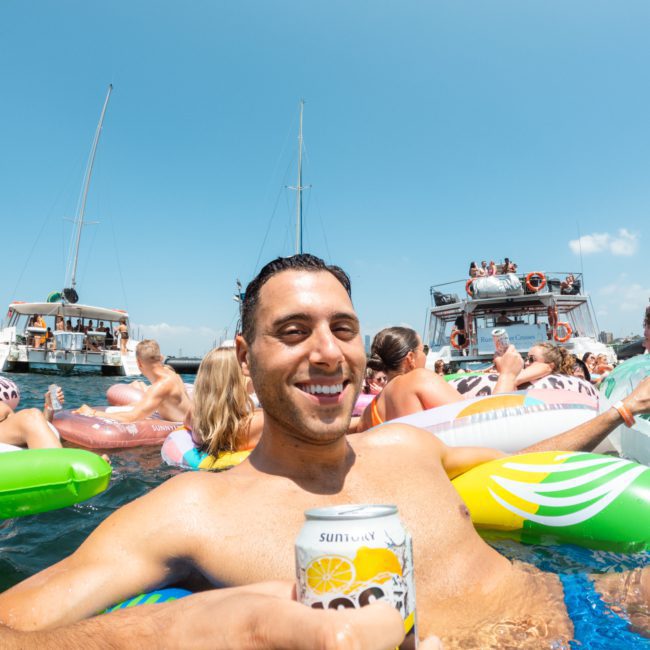 People relaxing on various inflatable floaties in water with boats in the background. A foreground person is smiling and holding a beverage can. The sky is clear and sunny, perfect for Corporate boat events Sydney.