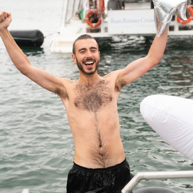 Man in black shorts stands in water with arms raised in celebration. A boat and other people are visible in the background, reminiscent of a lively catamaran party Sydney.
