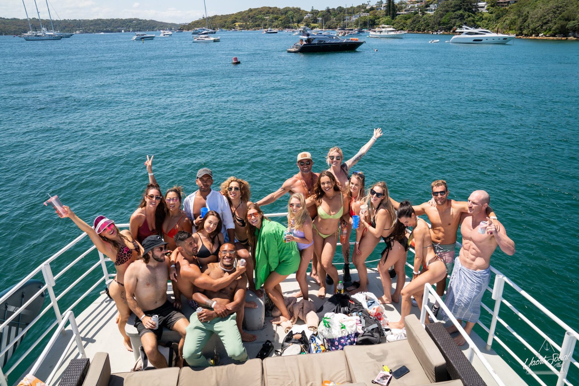 A large group of people poses on a boat deck, many in swimwear, with the ocean and other boats visible in the background under a clear sky. It's the perfect scene for a Sydney boat party hire.