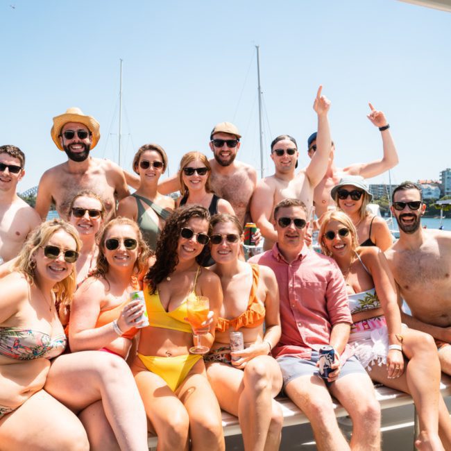 A group of 17 people in swimwear are gathered on a boat, smiling and posing for a photo with a waterfront and boats visible in the background, enjoying their private yacht charter Sydney Harbour.