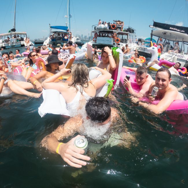 A group of people enjoying a sunny day on inflatable floaties in the water, surrounded by boats. Some are holding drinks and socializing. The environment appears lively and festive, perfect for those considering a catamaran party in Sydney.