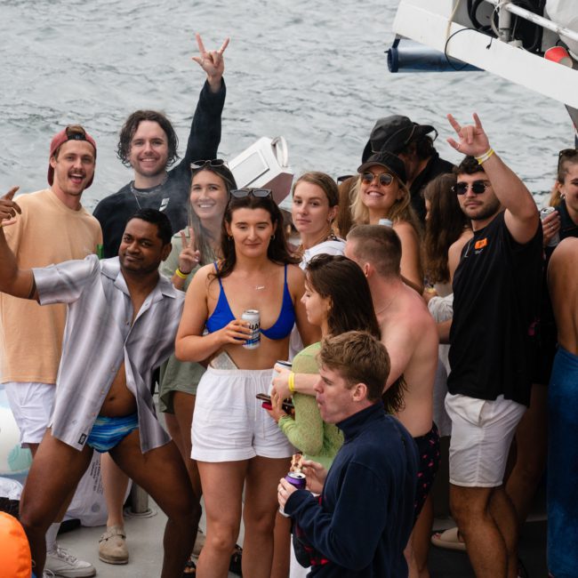 A group of people on a boat, some holding drinks and wearing swimwear, smiling and making gestures. The background shows water and parts of the boat, perfect for a catamaran party in Sydney.