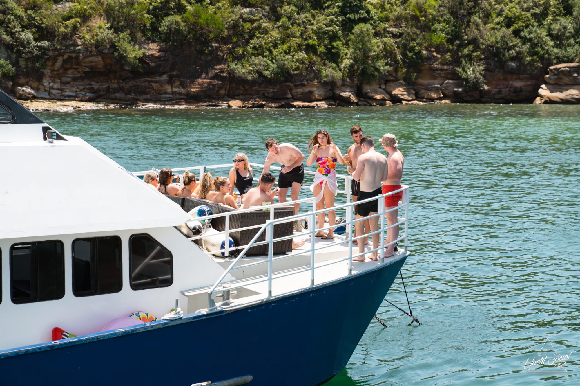 A group of people in swimsuits are gathered on the deck of a luxury yacht floating near a rocky shoreline with greenery.