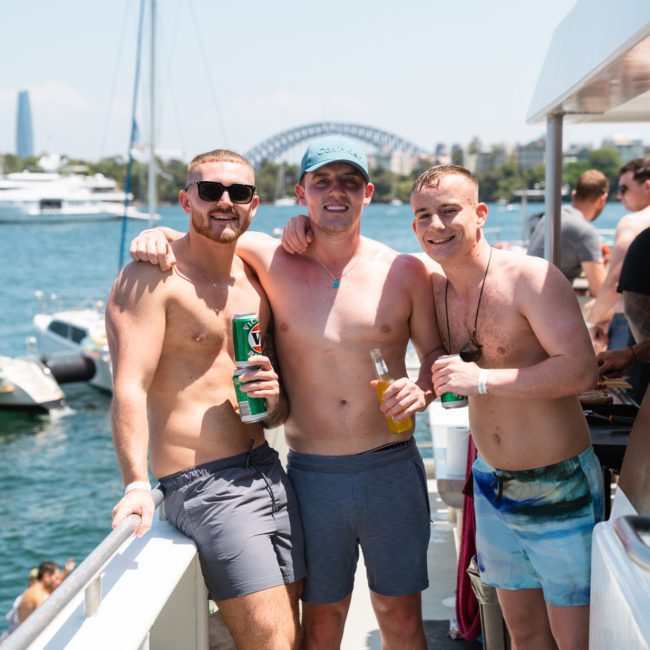 Three men in swimwear stand on a boat, smiling and holding drinks. A marina with boats and a bridge can be seen in the background, embodying the essence of Sydney boat party hire.