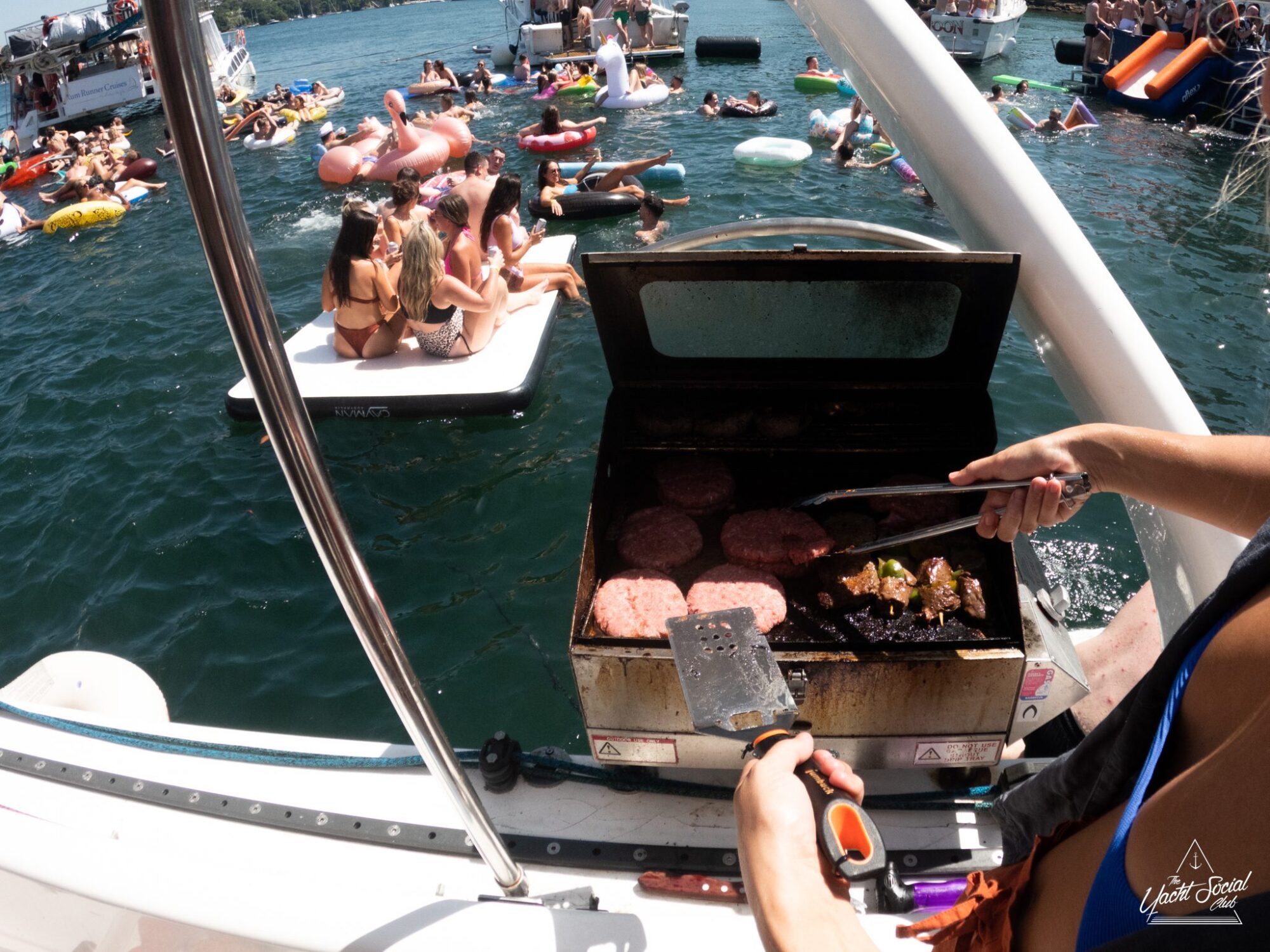 People are enjoying a pool party on floats and boats in a lake, while someone grills burgers and vegetables on a luxury yacht.