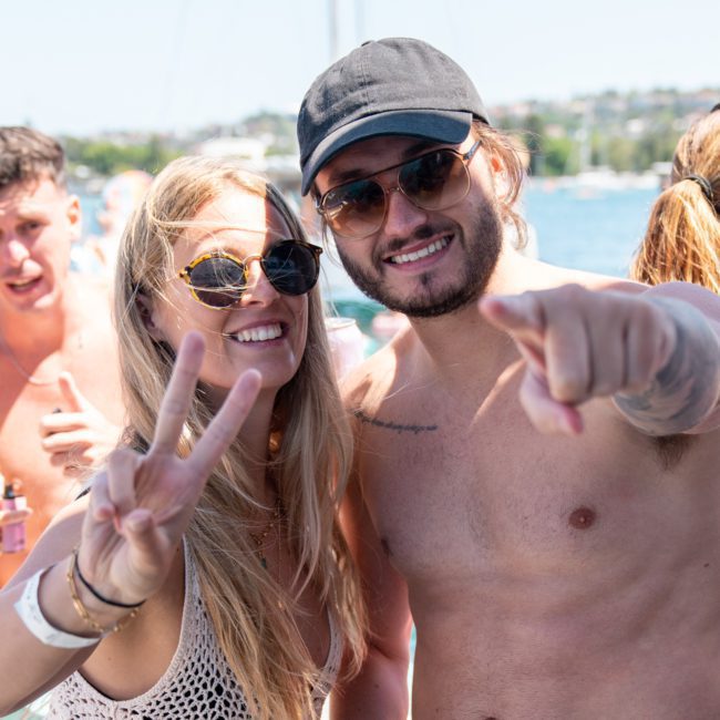 A woman and a man pose for a photo on a boat. The woman is flashing a peace sign and smiling, while the man points at the camera. They are wearing sunglasses and casual summer clothing, enjoying their luxury yacht hire in Sydney.