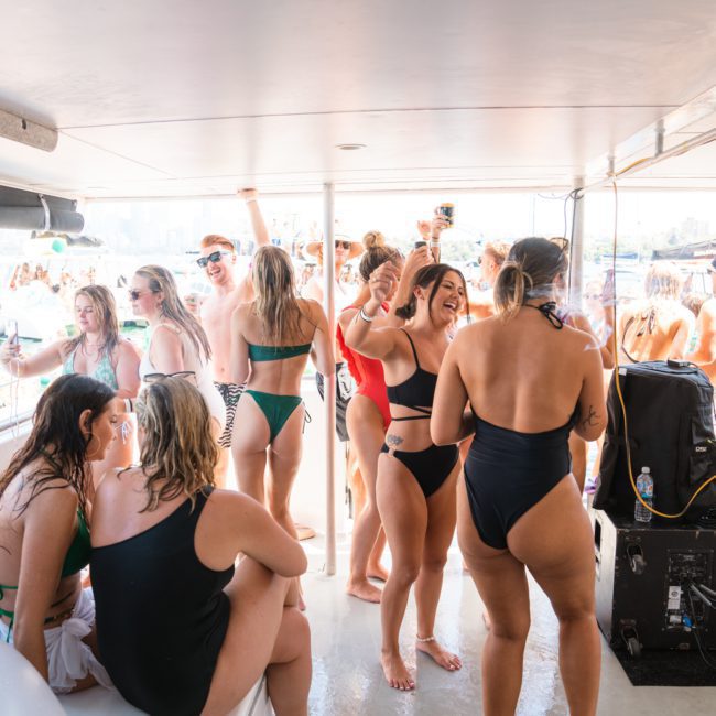 A group of people in swimwear are socializing and dancing on a private yacht charter in Sydney Harbour during the daytime. There is a sound system setup, and other boats are visible in the background.