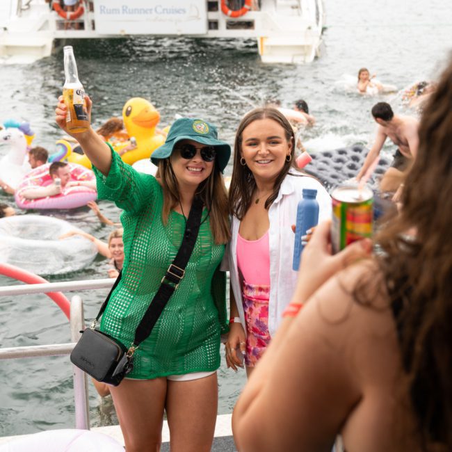 Two women in swimsuits and beachwear, one holding a beer bottle, pose for a photo in front of a crowded Catamaran party Sydney with people enjoying the water. The scene suggests a lively outdoor event or party.