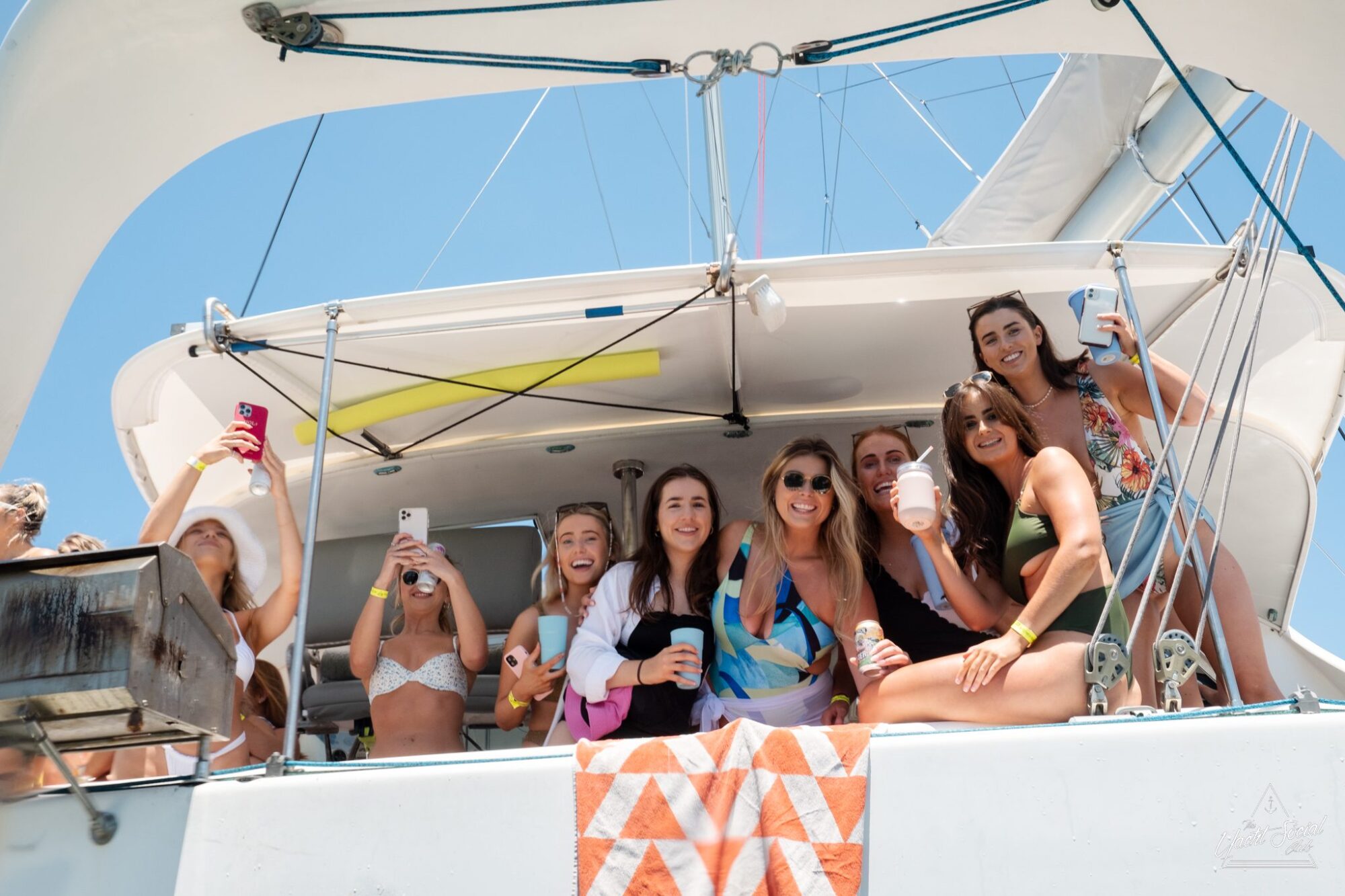 A group of smiling women on a boat holding drinks, with a towel draped on the railing. Some are taking photos, and it is a sunny day, perfect for a corporate boat event in Sydney Harbour.