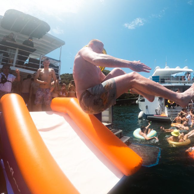 A man in swim trunks slides down an orange and white inflatable slide into the water from a boat. Other people in swimwear watch and swim nearby, enjoying a lively atmosphere. Perfect for those considering DJ boat hire Sydney or private yacht charter Sydney Harbour, this scene epitomizes summer fun.