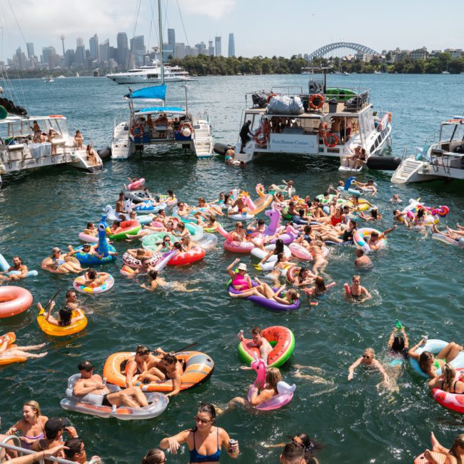 A group of people swimming and floating on colorful inflatables in the water, with several boats and a city skyline in the background, enjoying a Luxury yacht hire Sydney.