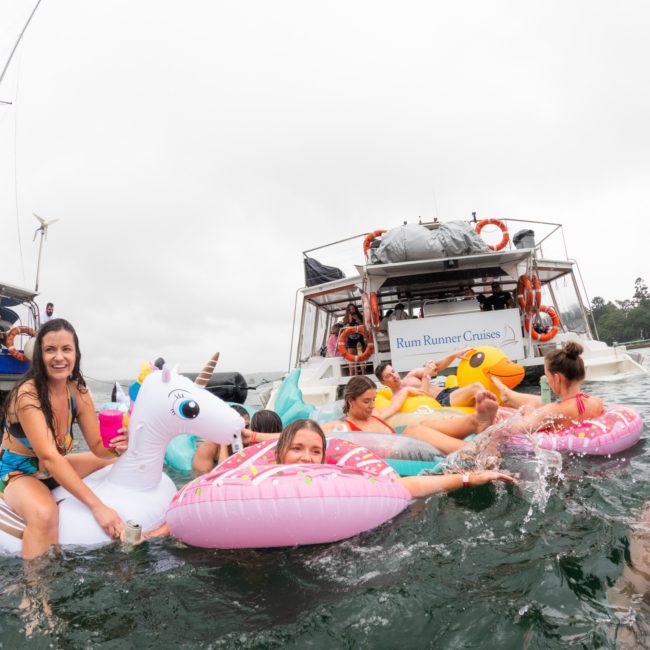 People enjoying a gathering in a body of water, floating on various inflatable pool toys near a boat labeled "Rum Runner Cruises." The scene exudes the festive vibe of a Catamaran party Sydney, creating an unforgettable experience.