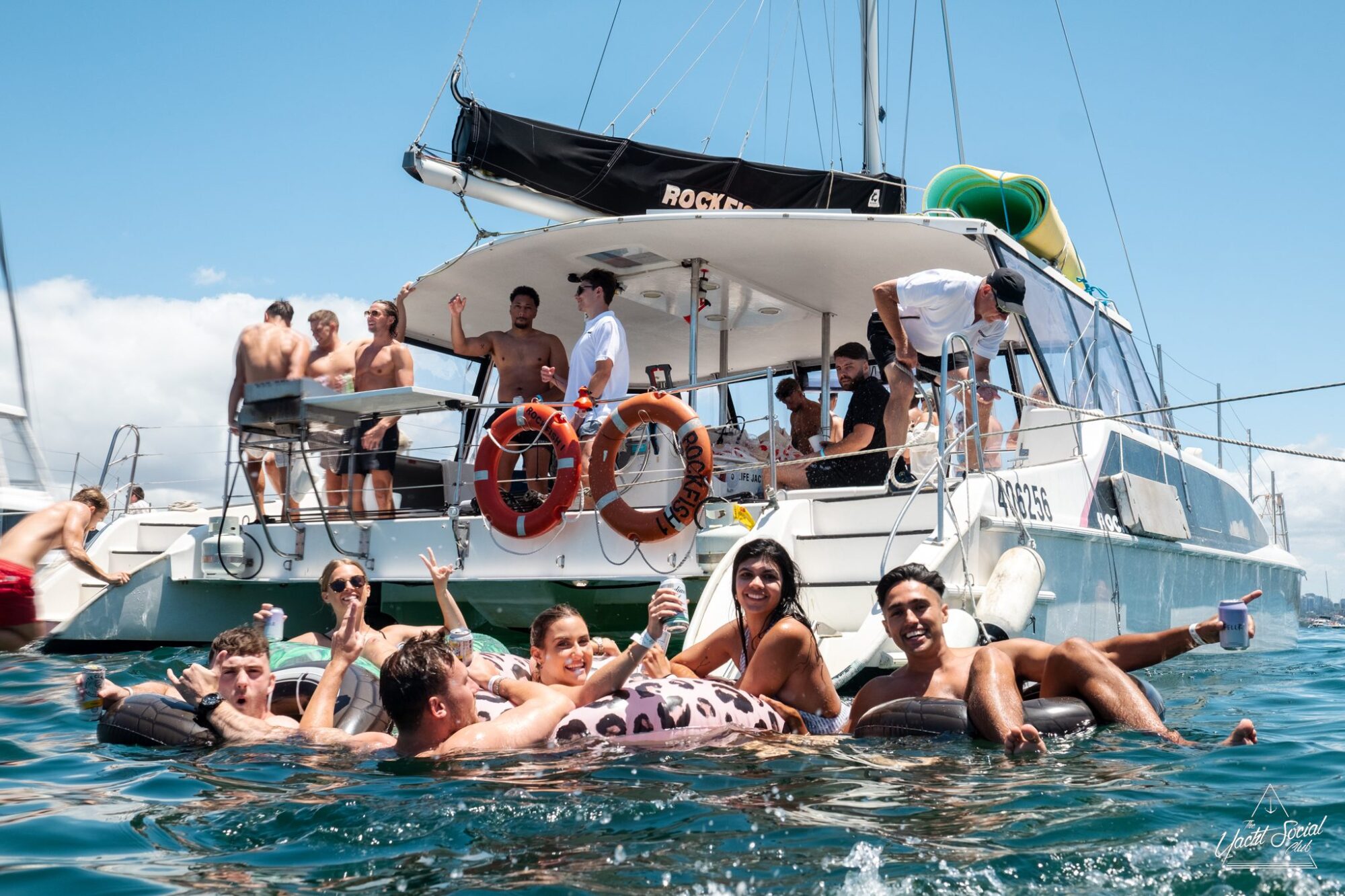 A group of people relax on and around a luxury yacht, some floating on inflatables in the water, enjoying a sunny day in Sydney Harbour.