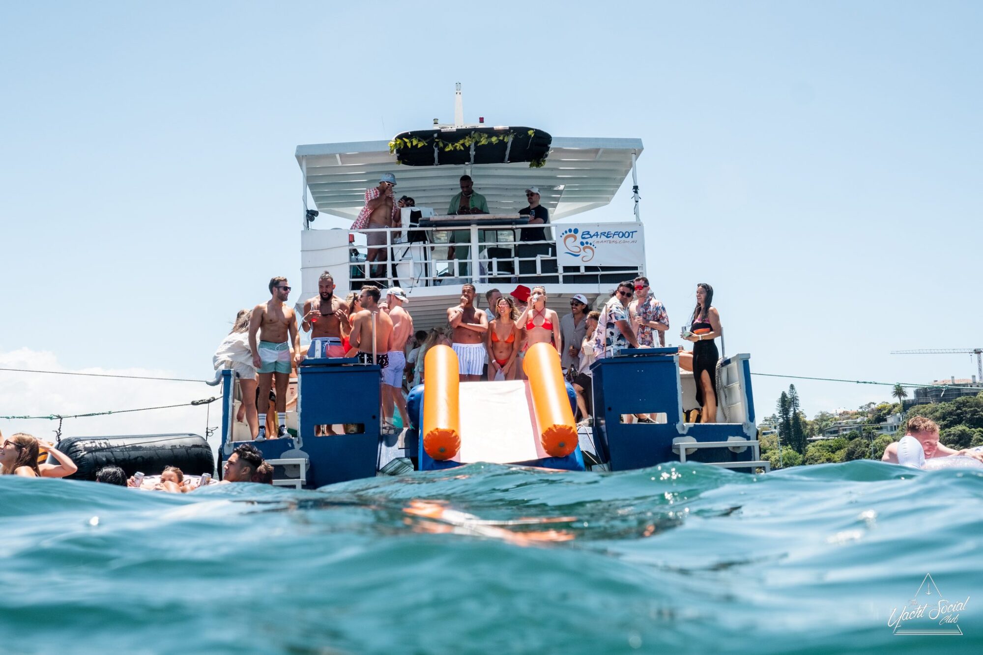 A group of people stand and relax on the deck of a luxury yacht at sea, with some individuals using large slides to enter the water. The boat, available for luxury yacht hire Sydney, has a logo that reads "Gamefoot.