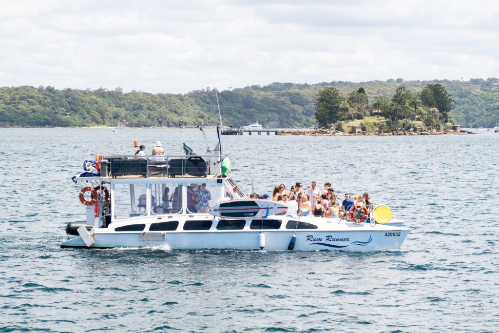 The "Rum Runner," a pristine white yacht, is carrying a lively group as it sails near a small, tree-covered island, epitomizing the elegance of luxury yacht hire in Sydney.