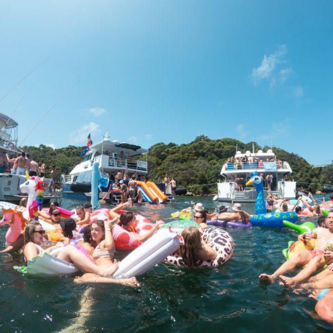 People relaxing on various inflatable floats in the water near anchored boats on a sunny day, enjoying a DJ boat hire Sydney experience.